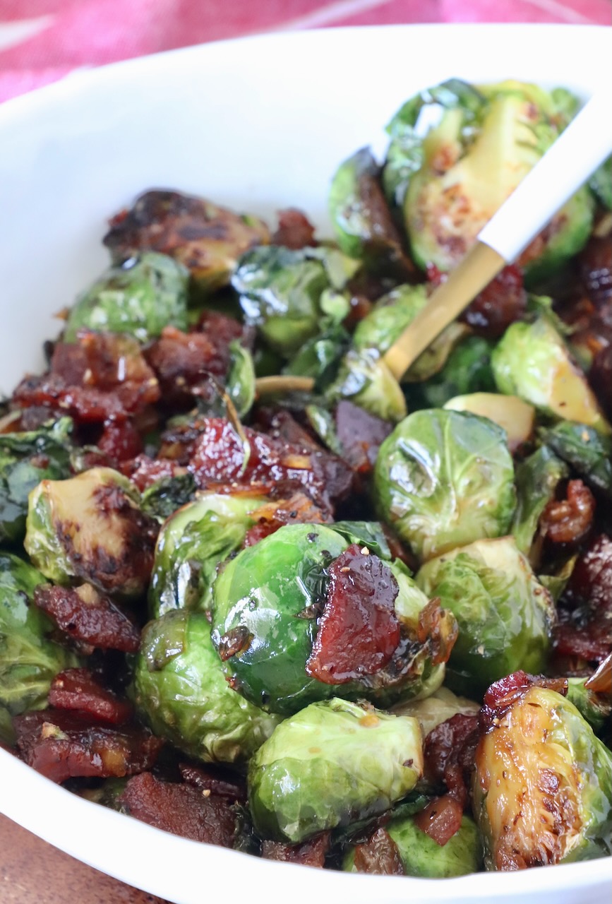 Caramelized Brussels sprouts with crispy bacon pieces in a white bowl. The sprouts are glazed, showing a glossy texture.