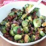 Caramelized Brussels sprouts with crispy bacon pieces in a white bowl with a serving spoon.