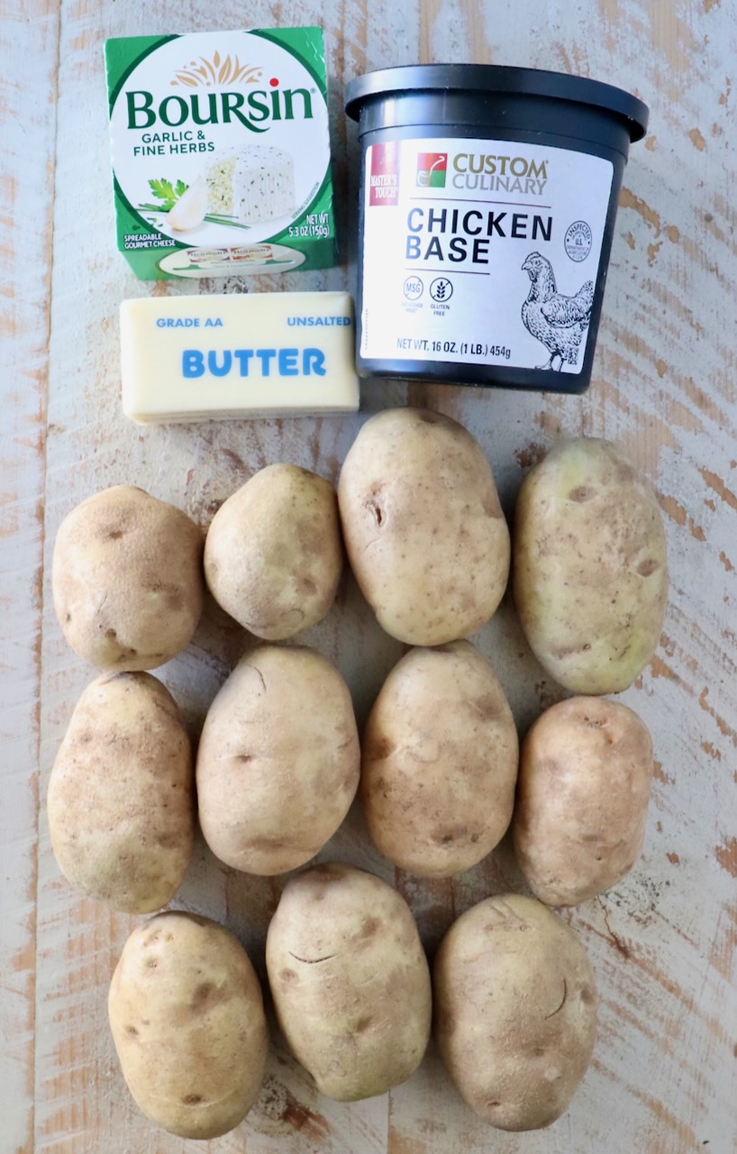 Twelve potatoes arranged in three rows, accompanied by a box of Boursin cheese, a container of chicken base, and a stick of unsalted butter on a wooden surface.