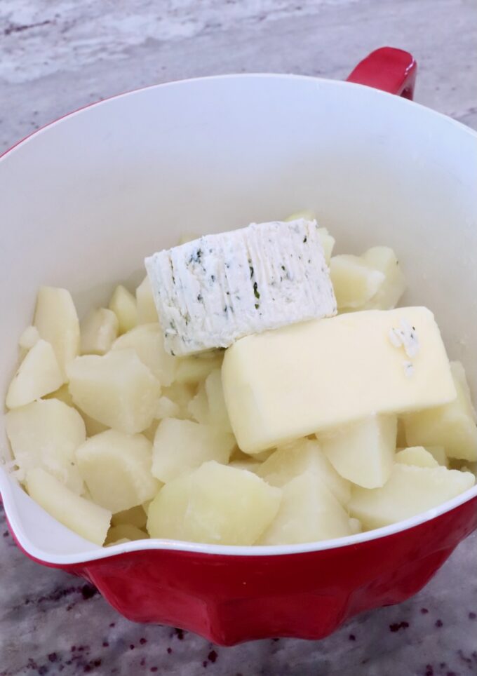 cubed potatoes in a red bowl with a stick of butter and a round of herbed cheese on top