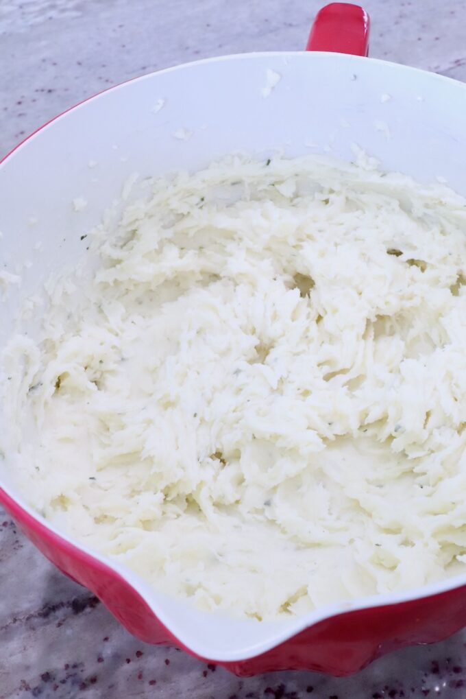Mashed potatoes in a red and white bowl on a speckled countertop. The texture appears creamy and smooth