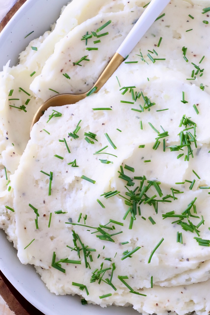 Creamy mashed potatoes garnished with fresh chopped chives in a white bowl. A gold and white spoon is dipped in the potatoes.