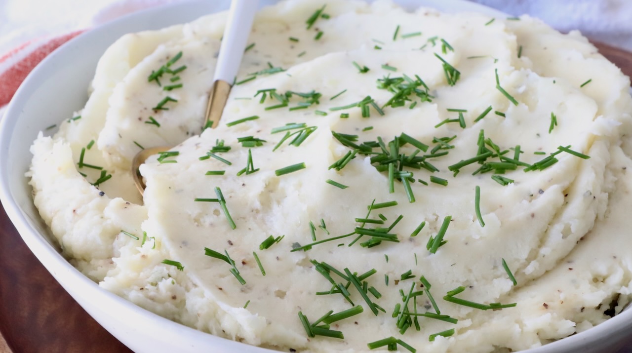 A white bowl filled with creamy mashed potatoes garnished with fresh green chives. A golden spoon rests in the dish.
