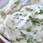 Creamy mashed potatoes garnished with fresh chopped chives in a white bowl. A gold and white spoon is dipped in the potatoes.