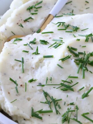 Creamy mashed potatoes garnished with fresh chopped chives in a white bowl. A gold and white spoon is dipped in the potatoes.
