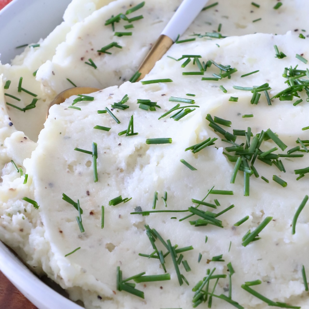 Creamy mashed potatoes garnished with fresh chopped chives in a white bowl. A gold and white spoon is dipped in the potatoes.