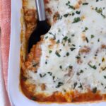Close-up of butternut squash baked ravioli in a white dish. It features a golden-brown cheese topping sprinkled with herbs, with a serving spatula on the side.