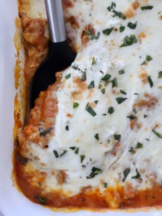 Close-up of butternut squash baked ravioli in a white dish. It features a golden-brown cheese topping sprinkled with herbs, with a serving spatula on the side.