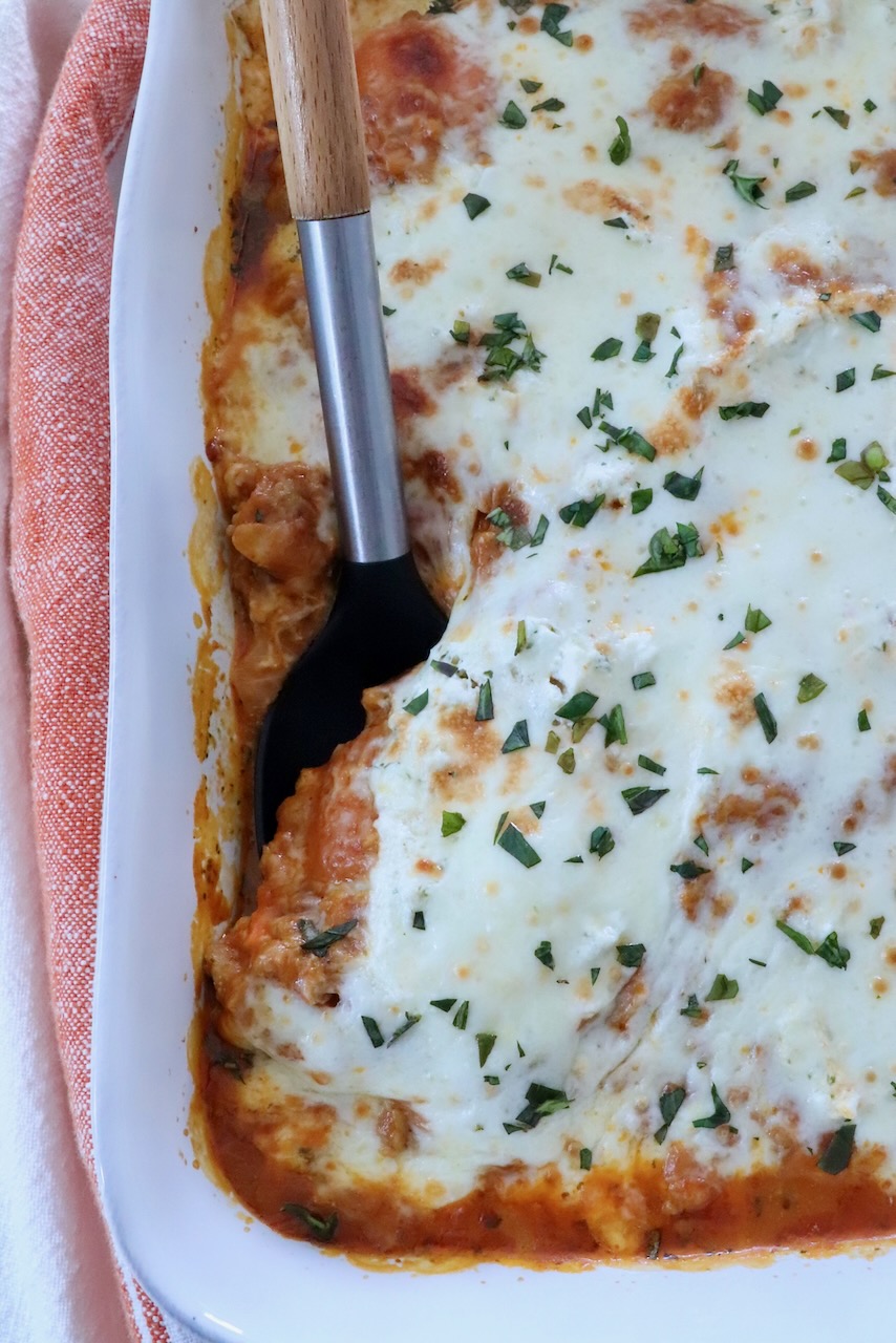 Close-up of butternut squash baked ravioli in a white baking dish, featuring melted cheese sprinkled with herbs. A serving has been removed with a black spatula.