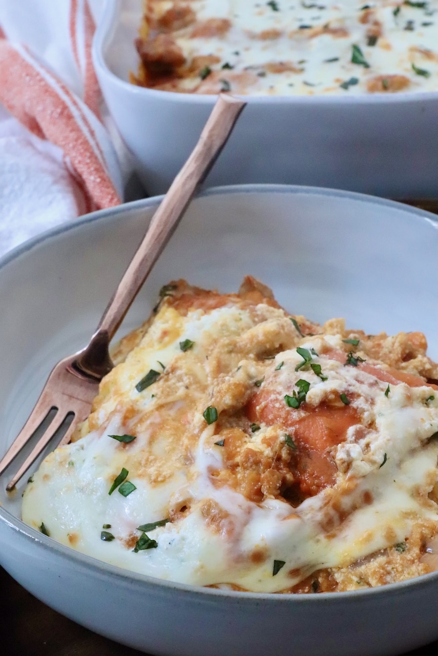 A close-up of a creamy, cheesy pasta bake in a bowl with a fork, garnished with fresh herbs. In the background, a baking dish of the same pasta.