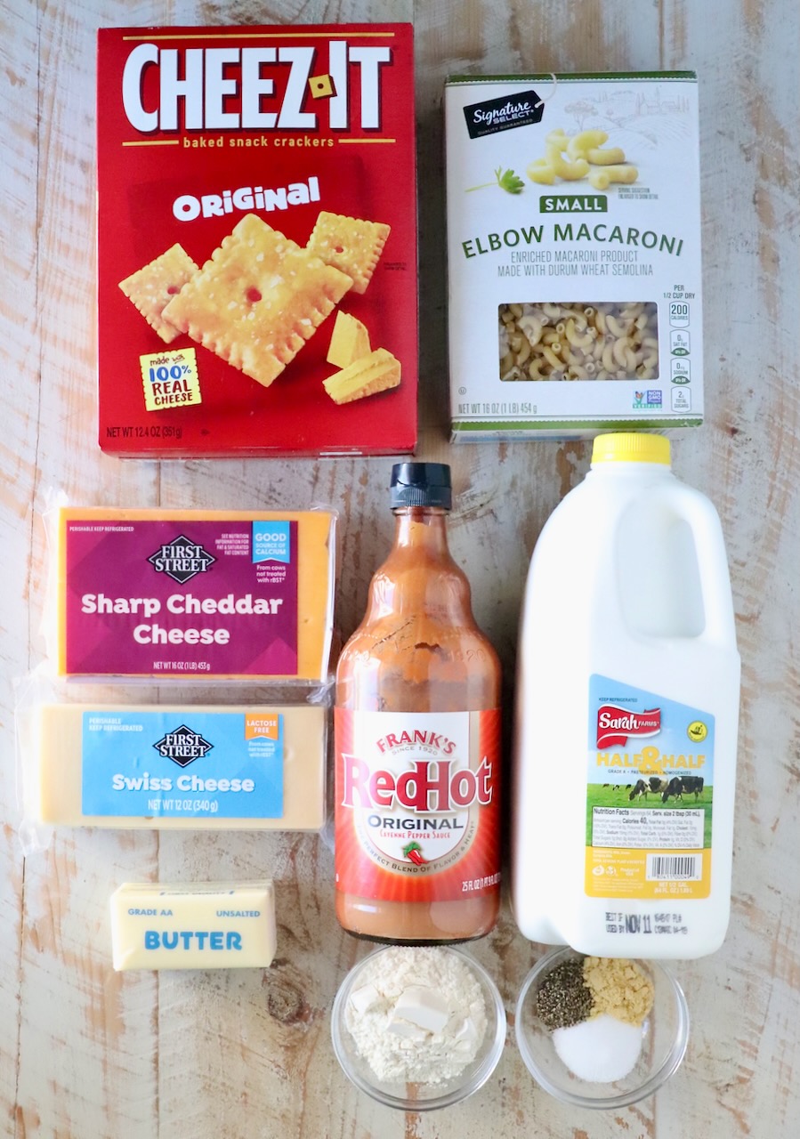Assorted ingredients for mac and cheese on a wooden surface, including a box of Cheez-It crackers, elbow macaroni, cheddar and Swiss cheeses, hot sauce, milk, butter, flour, and spices.