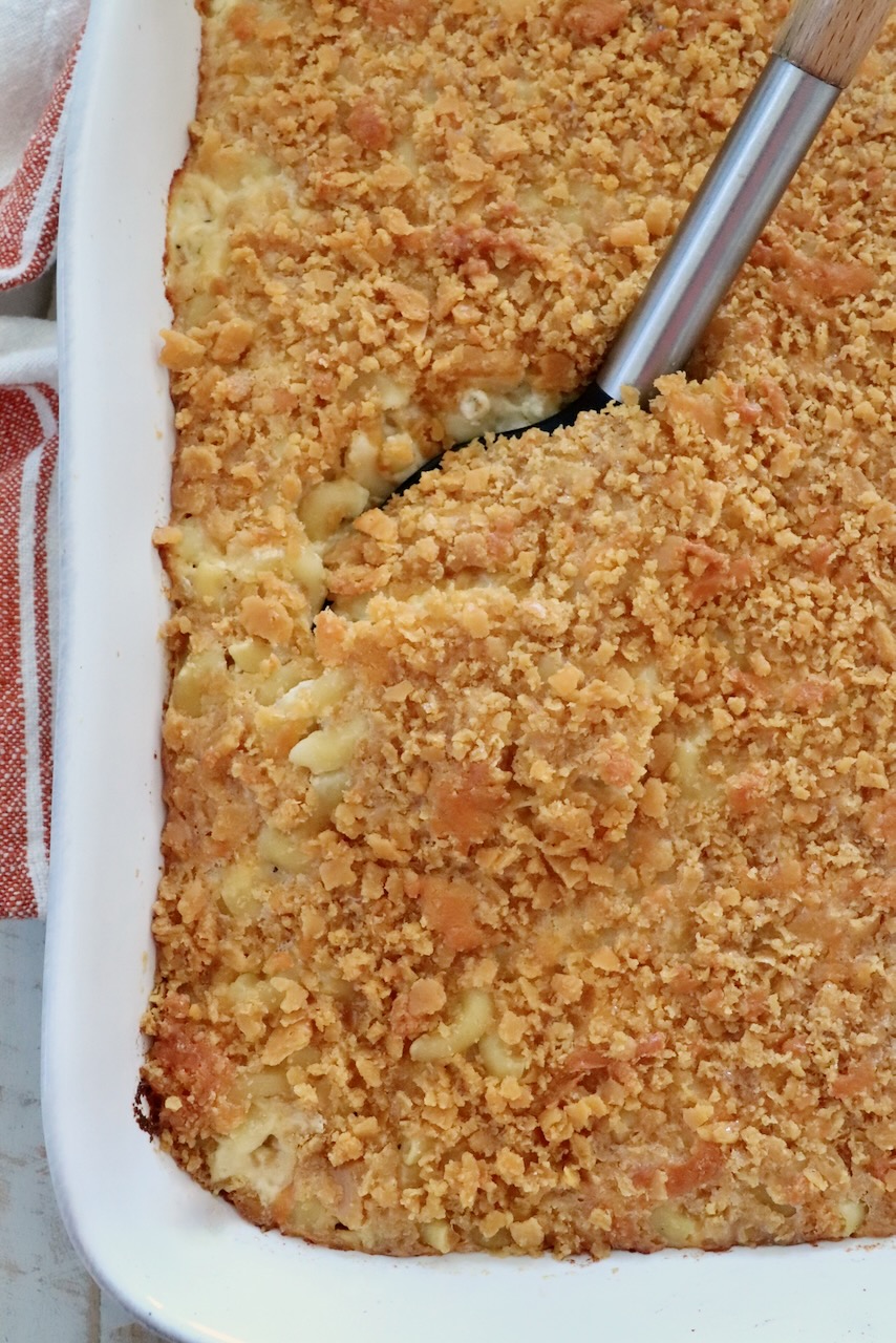 Golden-brown baked macaroni and cheese with a crispy cracker topping in a white casserole dish. A serving spoon is lifting a portion, revealing creamy pasta underneath.