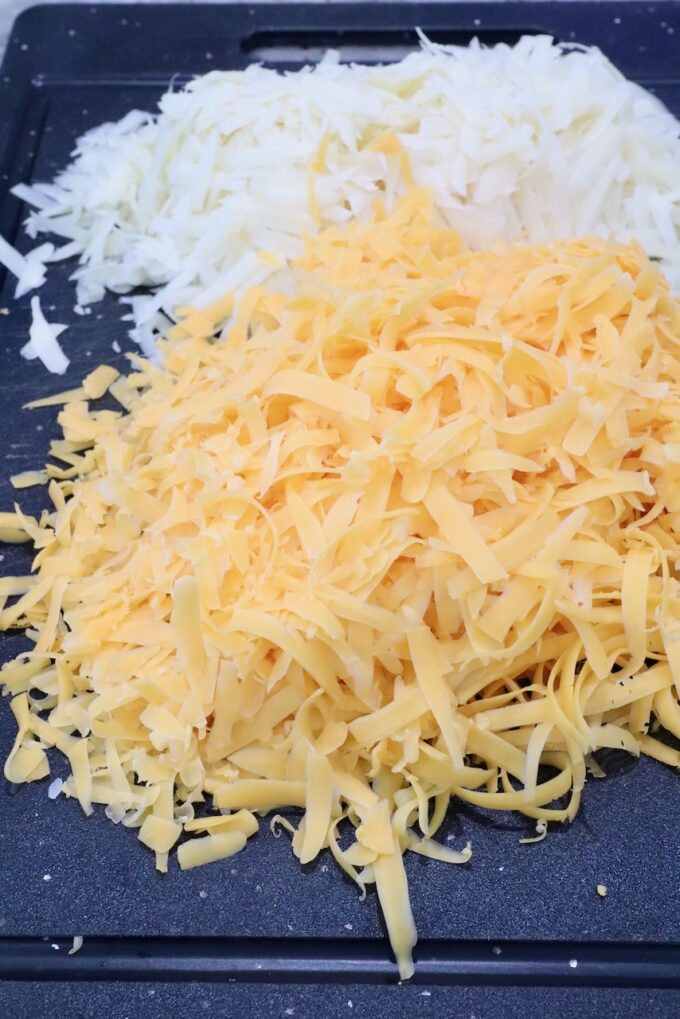 A pile of finely grated swiss cheese and a separate pile of cheddar cheese are on a black cutting board