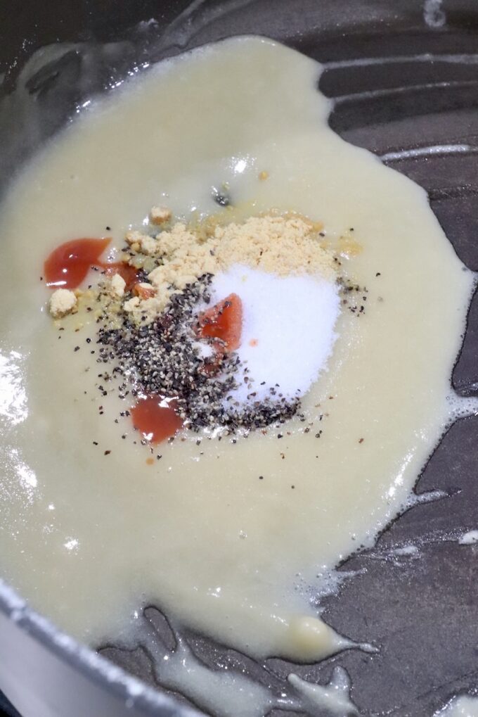 A close-up of a skillet shows a roux with added spices, including, black pepper, red hot sauce, and a teaspoon of salt