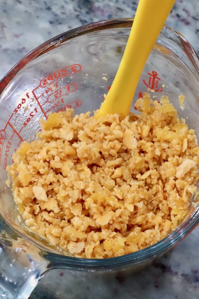 A clear measuring cup filled with crumbled Cheez-Its sits on a marble countertop. A bright yellow spoon is partially immersed in the crushed crackers.