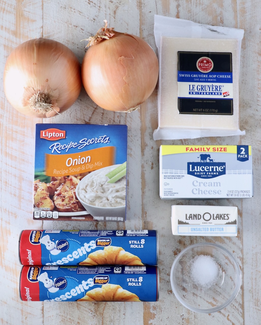 Ingredients on a wooden surface include two onions, Gruyère cheese, onion soup mix, cream cheese, crescent roll tubes, butter, and salt.