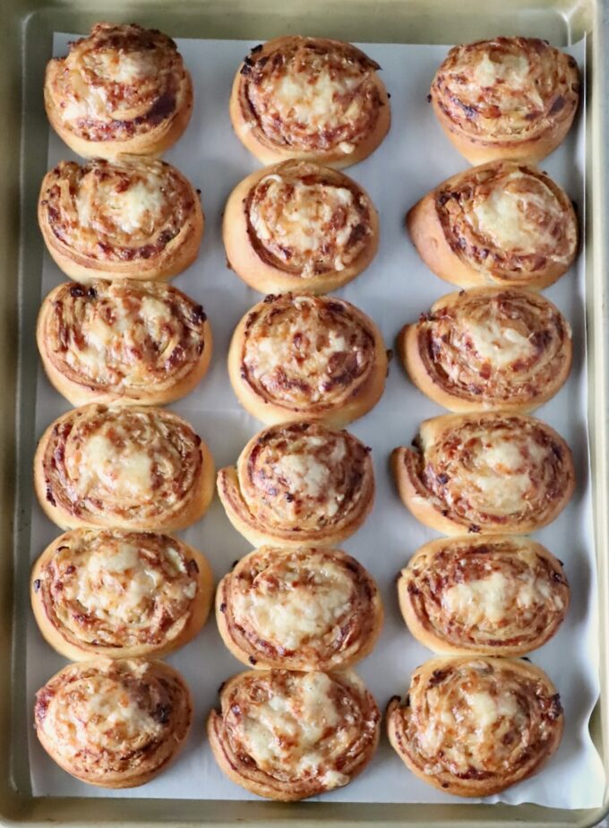 Eighteen golden-brown cheese and caramelized onion rolls on a baking sheet, evenly arranged in three rows.