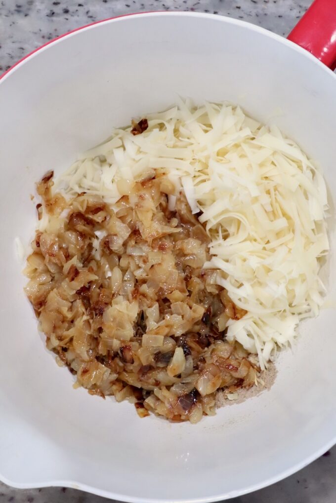 Chopped caramelized onions and shredded cheese in a white mixing bowl on a speckled countertop