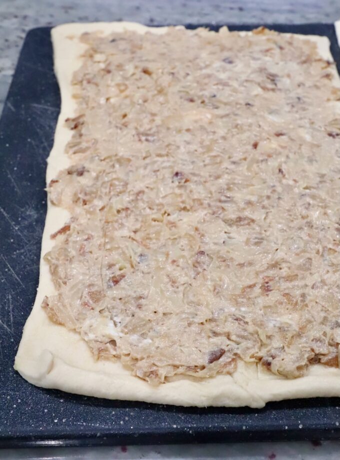 Rectangular dough on a black cutting board, topped with a creamy mixture of caramelized onions, covering the entire surface. 