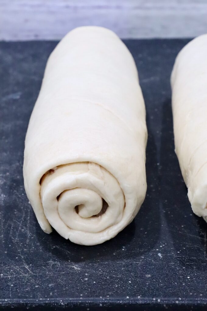 Two neatly rolled croissant dough logs rest side by side on a dark, floured surface, ready for baking.