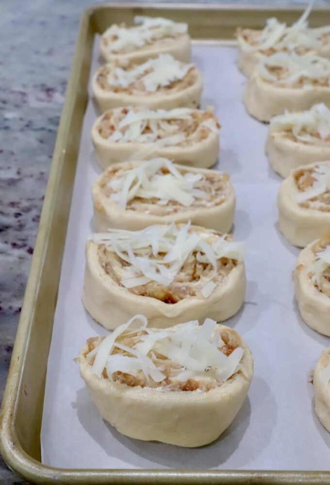 Close-up of unbaked caramelized onion rolls on a parchment-lined baking sheet. Each roll is topped with shredded cheese.