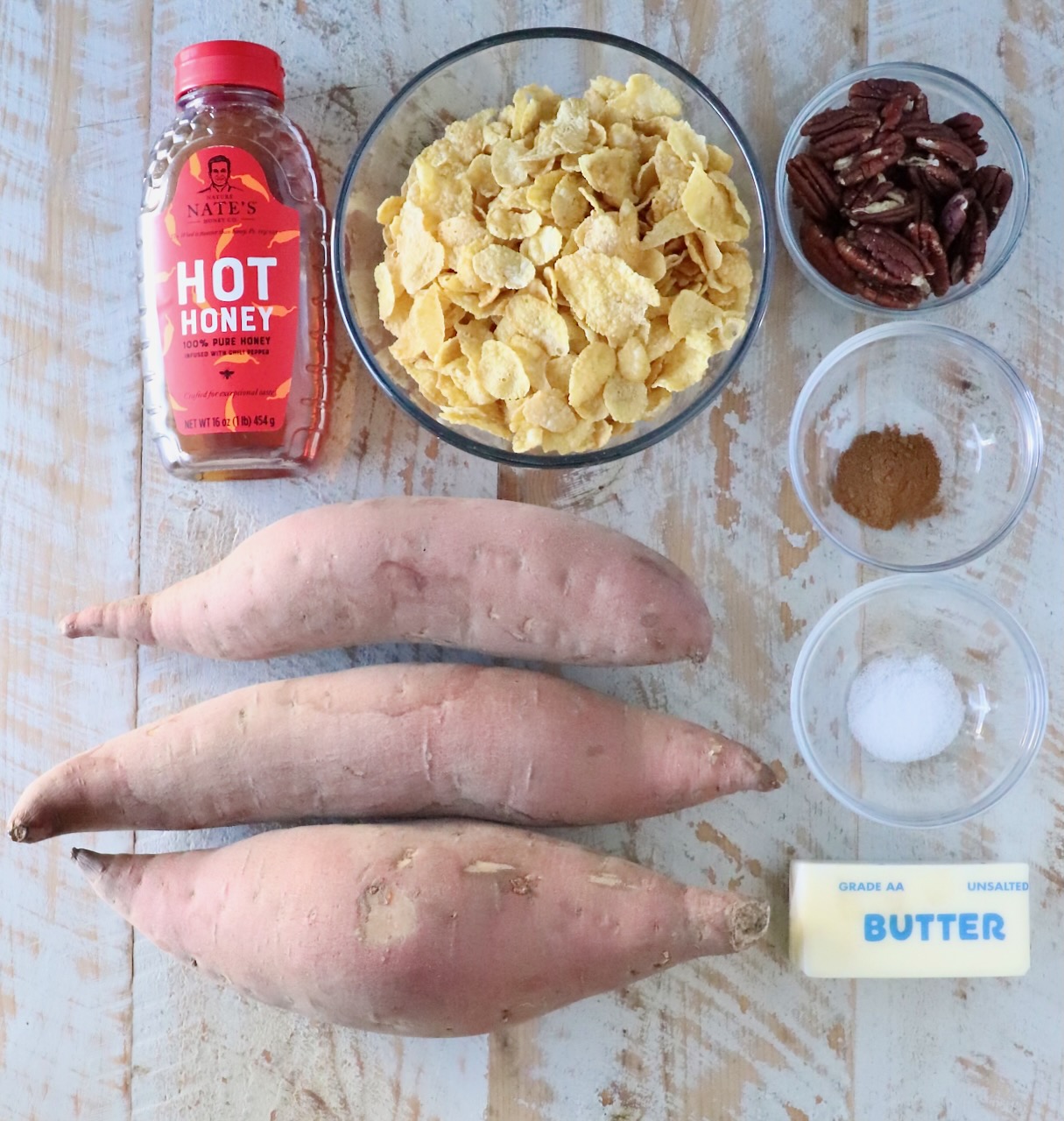 Ingredients for a sweet potato dish on a wooden surface: three raw sweet potatoes, a bottle of hot honey, cornflakes in a bowl, pecans, cinnamon, salt, and a stick of butter.