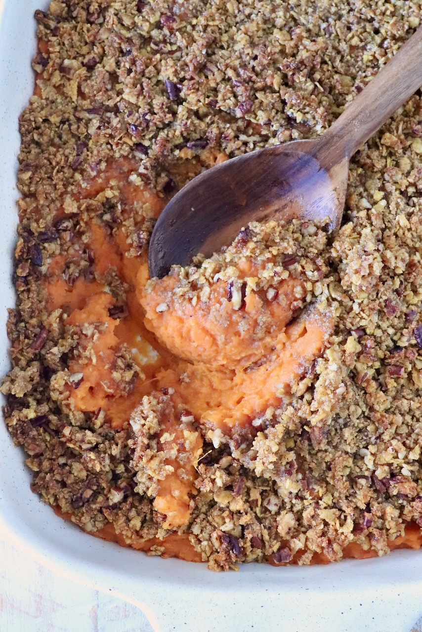 Close-up of a sweet potato casserole with a crunchy pecan topping and a wooden spoon scooping the orange sweet potato filling out from the dish