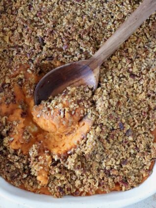 Close-up of a sweet potato casserole with a crunchy pecan topping and a wooden spoon scooping the orange sweet potato filling out from the dish