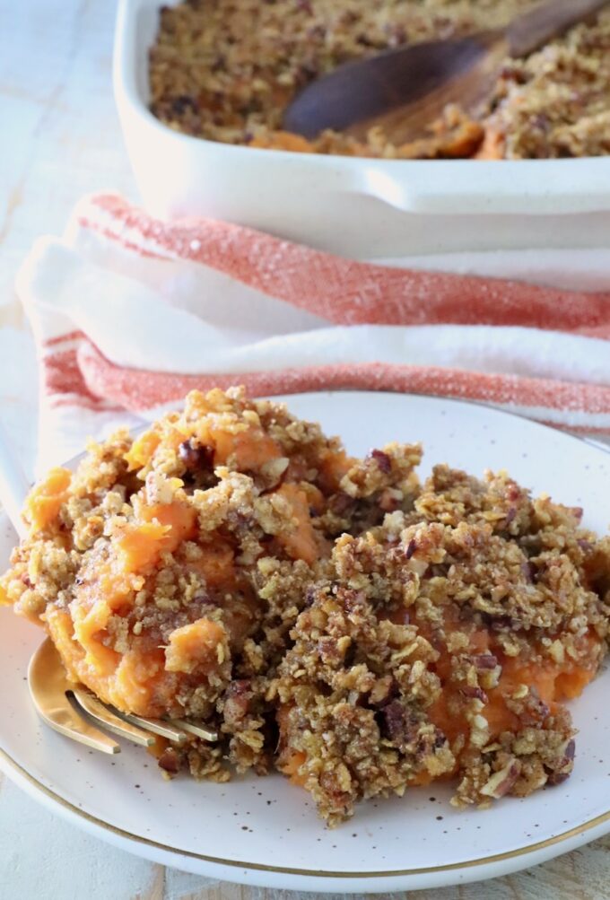 Plate of sweet potato casserole topped with a crispy pecan-crumb layer. A serving spoon lies nearby; a baking dish and towel are in the background.