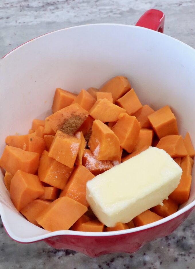 Cubed sweet potatoes with a stick of butter and sprinkled with spices, including salt and cinnamon, in a red mixing bowl on a marble counter.