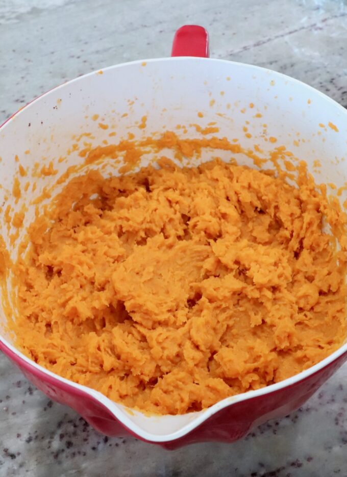 A red mixing bowl filled with smooth, bright orange mashed sweet potatoes sits on a speckled countertop