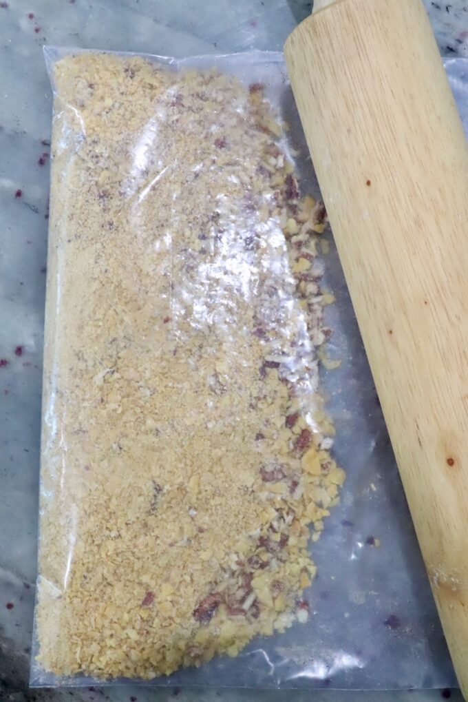 A plastic bag filled with crushed corn flakes and pecans lies on a marble surface next to a wooden rolling pin.