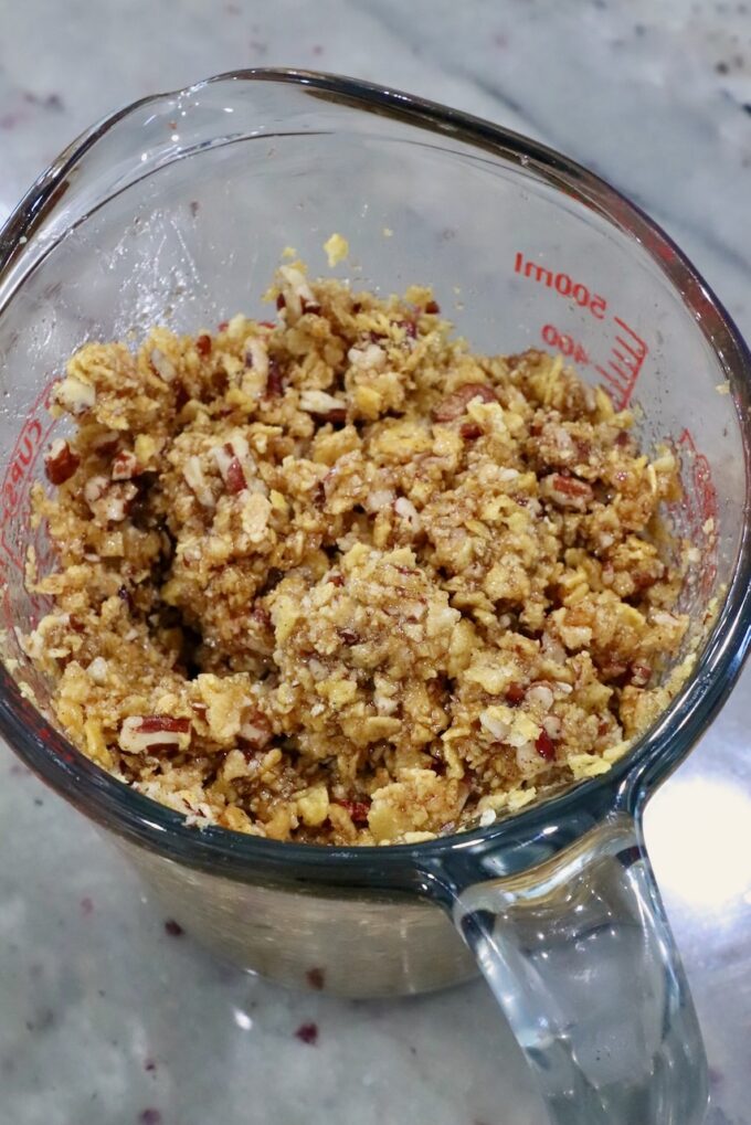 A glass measuring cup filled with a crumbly mix of corn flakes and chopped nuts sits on a speckled granite countertop