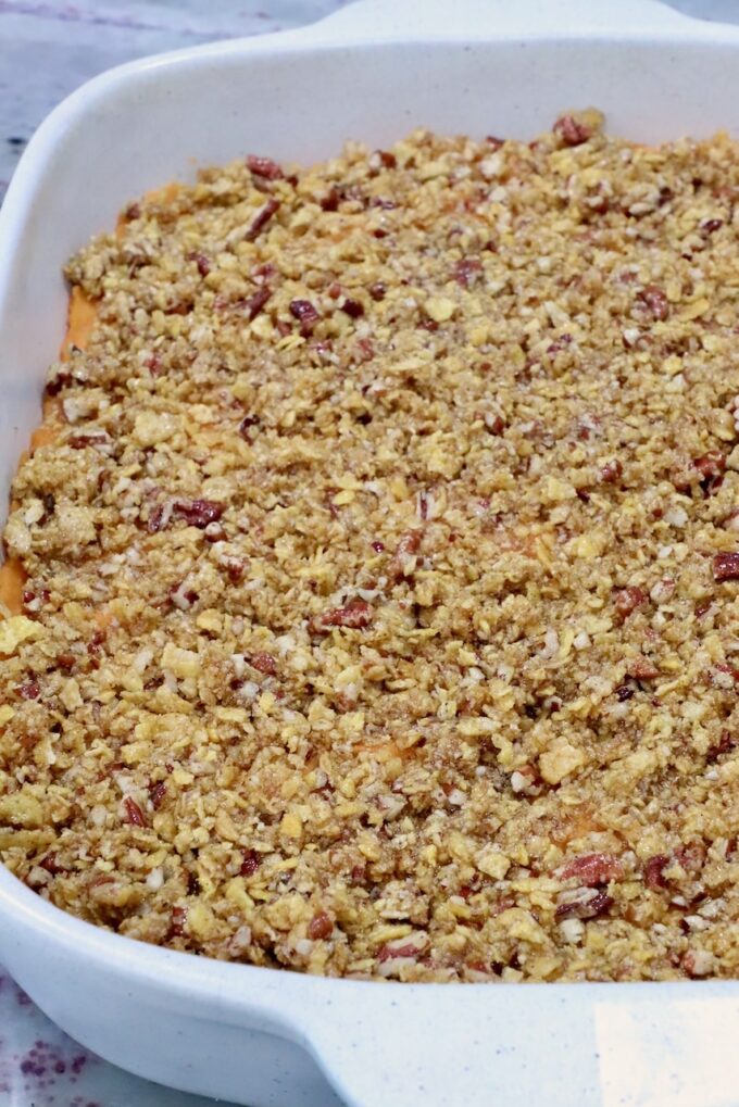 A white ceramic dish filled with a golden-brown, crumbly topping covering a sweet potato casserole, with visible pecan pieces throughout