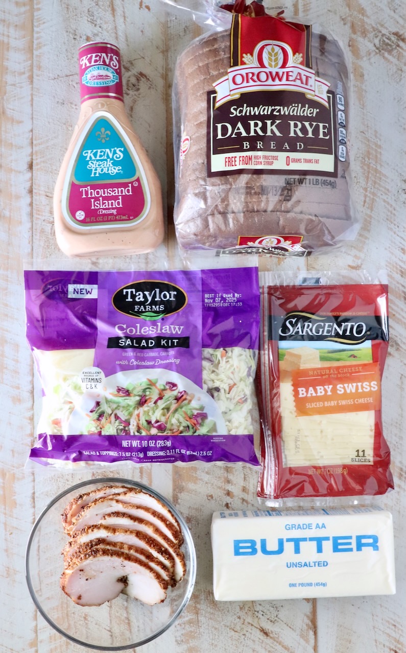 Packaged ingredients on a wooden table include Thousand Island dressing, dark rye bread, coleslaw kit, baby Swiss cheese, sliced cooked turkey, and butter.