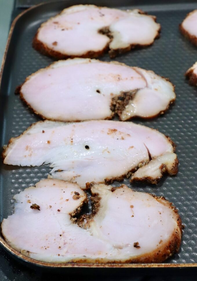 Slices of roasted turkey breast with a browned seasoning crust are laid out on a textured skillet.
