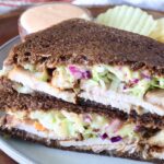 Close-up of a sandwich with dark rye bread filled with sliced turkey, creamy slaw, and sauce. Served on a plate with wavy potato chips and a dipping sauce.