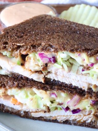 Close-up of a sandwich with dark rye bread filled with sliced turkey, creamy slaw, and sauce. Served on a plate with wavy potato chips and a dipping sauce.