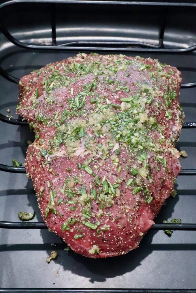 Raw seasoned beef roast covered in chopped herbs and spices, resting in a roasting pan. 