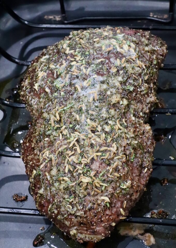 A seasoned roast beef resting on a wire rack, topped with herbs and spices, in a dark roasting pan