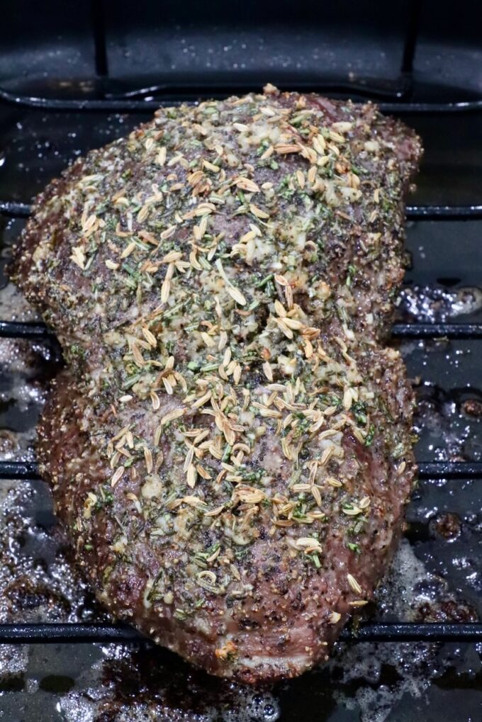 A seasoned roast beef resting on a wire rack, topped with herbs and spices, in a dark roasting pan