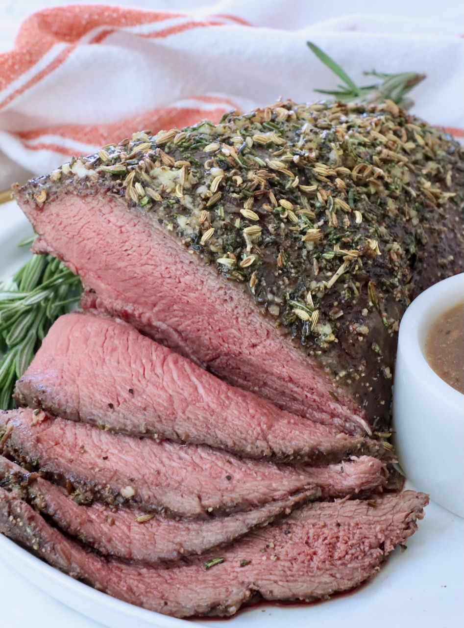 Roast beef topped with herbs and spices, sliced to reveal a juicy pink interior. Garnished with rosemary and served on a white plate with gravy.