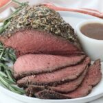 Roast beef topped with herbs and spices, sliced to reveal a juicy pink interior. Garnished with rosemary and served on a white plate with gravy.