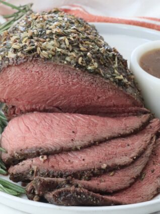 Roast beef topped with herbs and spices, sliced to reveal a juicy pink interior. Garnished with rosemary and served on a white plate with gravy.