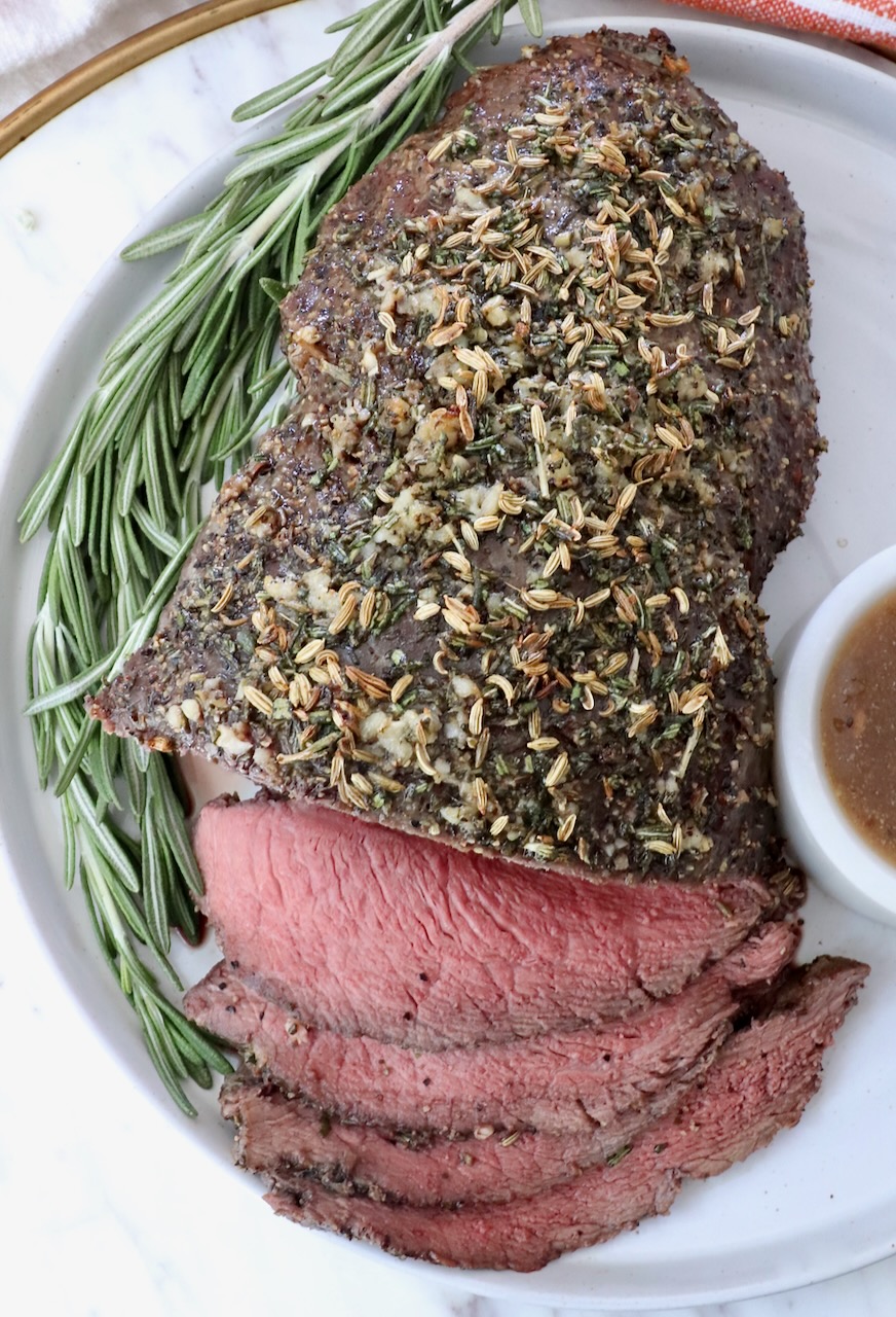 Roasted beef topped with herbs, sliced on a white plate with a sprig of rosemary. A small bowl of gravy sits on the side. 