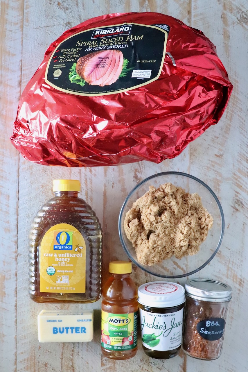 A collection of ingredients for ham on a wooden surface. Included are a large wrapped ham, brown sugar, honey, apple juice, butter, jam, and seasoning.