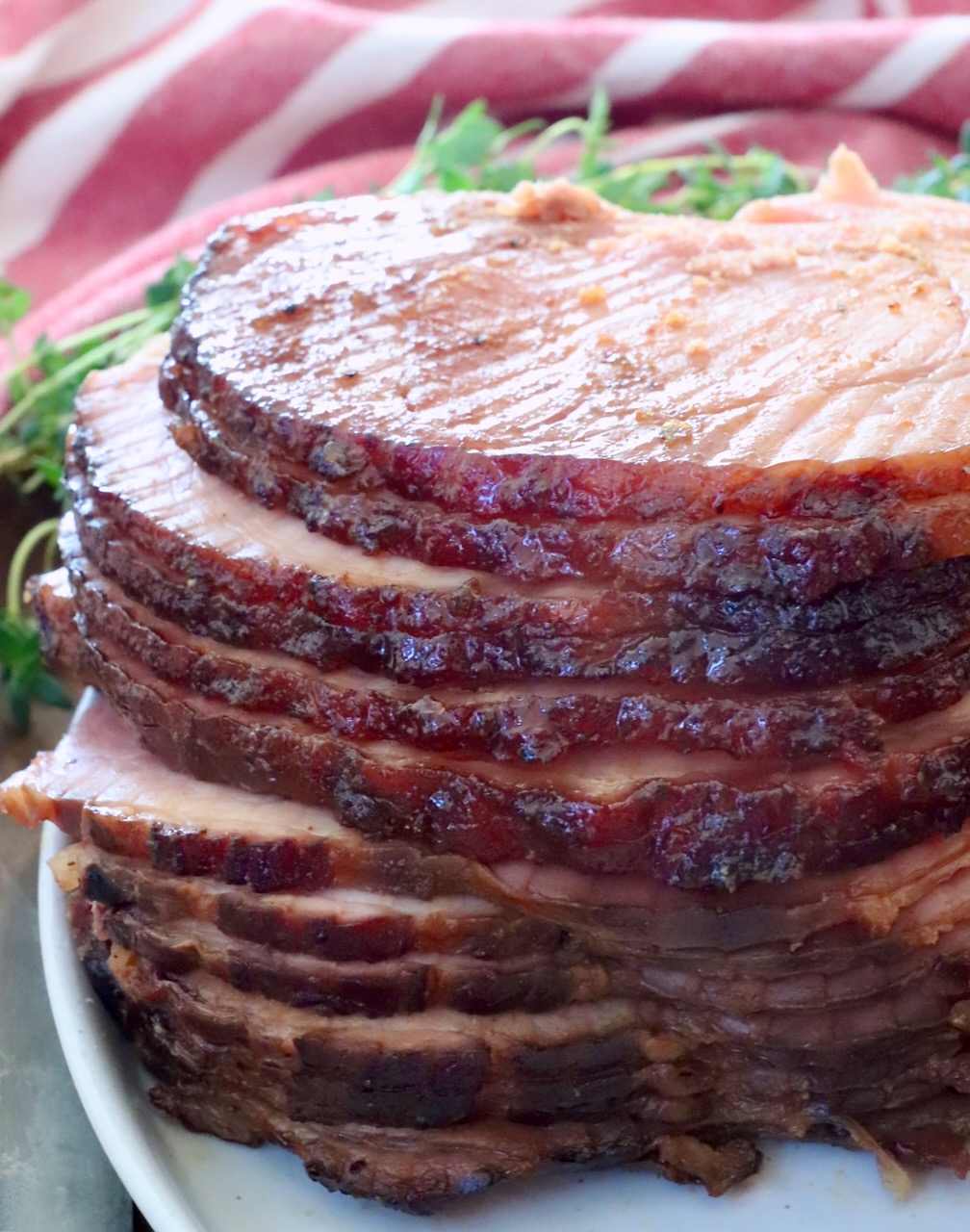 Sliced spiral ham glazed to a shiny brown, stacked on a white plate. Fresh herbs in the background and a red-striped cloth