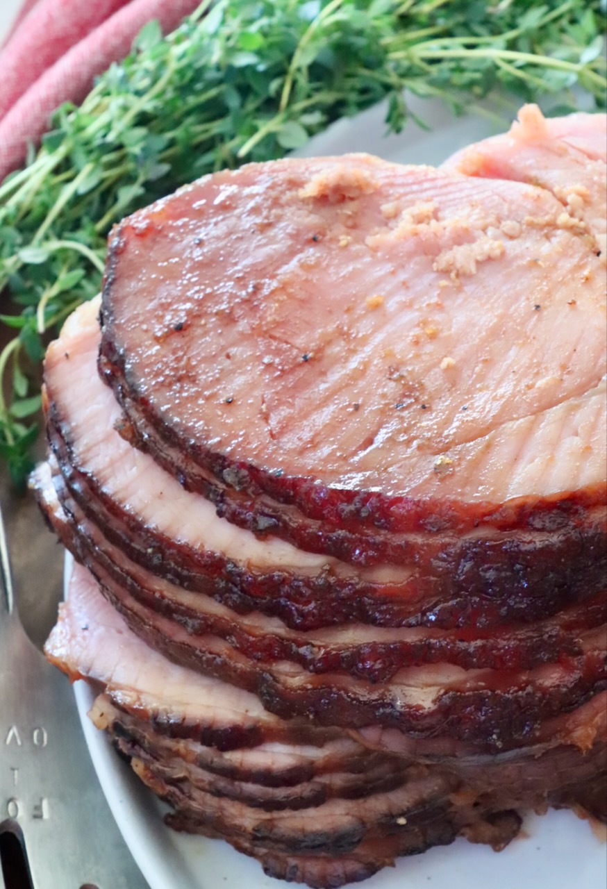Close-up of a glazed, spiral-cut ham on a white platter, showcasing juicy, tender slices. Fresh herbs and a red cloth are pictured in the background.