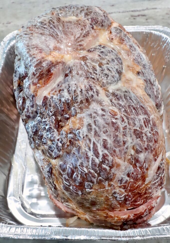 Cooked ham covered with melted butter, placed in a shiny aluminum tray. 