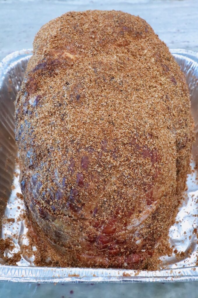 A large, seasoned ham covered in a brown sugar spice rub sits in an aluminum pan on a gray countertop, ready for cooking.